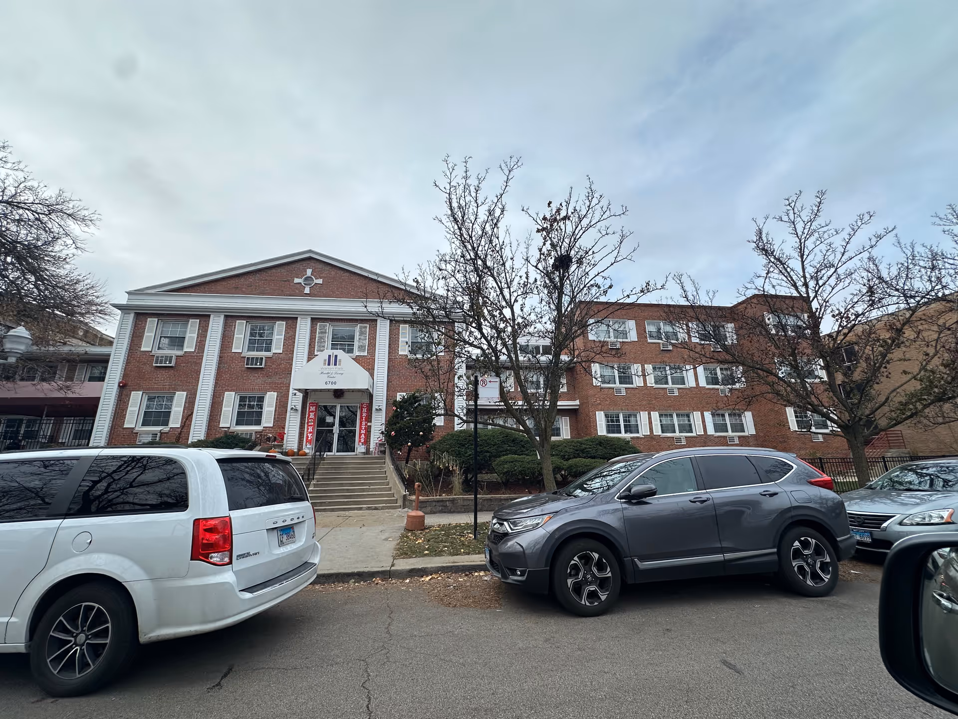 Exterior view of a three-story brick building with white window shutters and a white entrance canopy displaying the name Warren Park Health & Living Center. There are several parked cars in front of the building and leafless trees along the sidewalk.