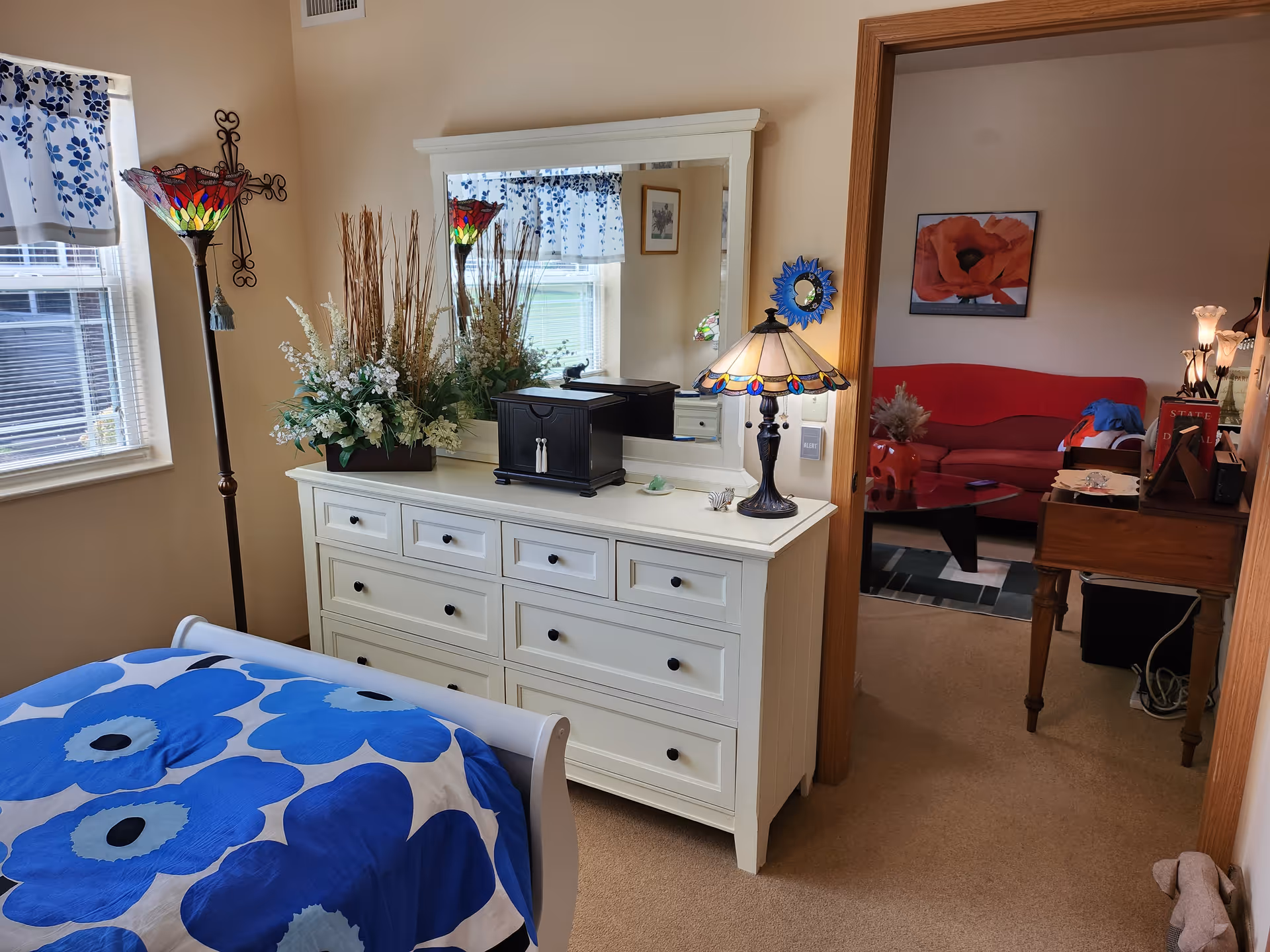 A cozy bedroom with a white dresser topped with a large mirror, a decorative lamp, and a flower arrangement. The bed has a blue and white floral patterned blanket. A window with a floral valance lets in natural light. Through an open doorway, a living room with a red couch, a coffee table, and wall art is visible.