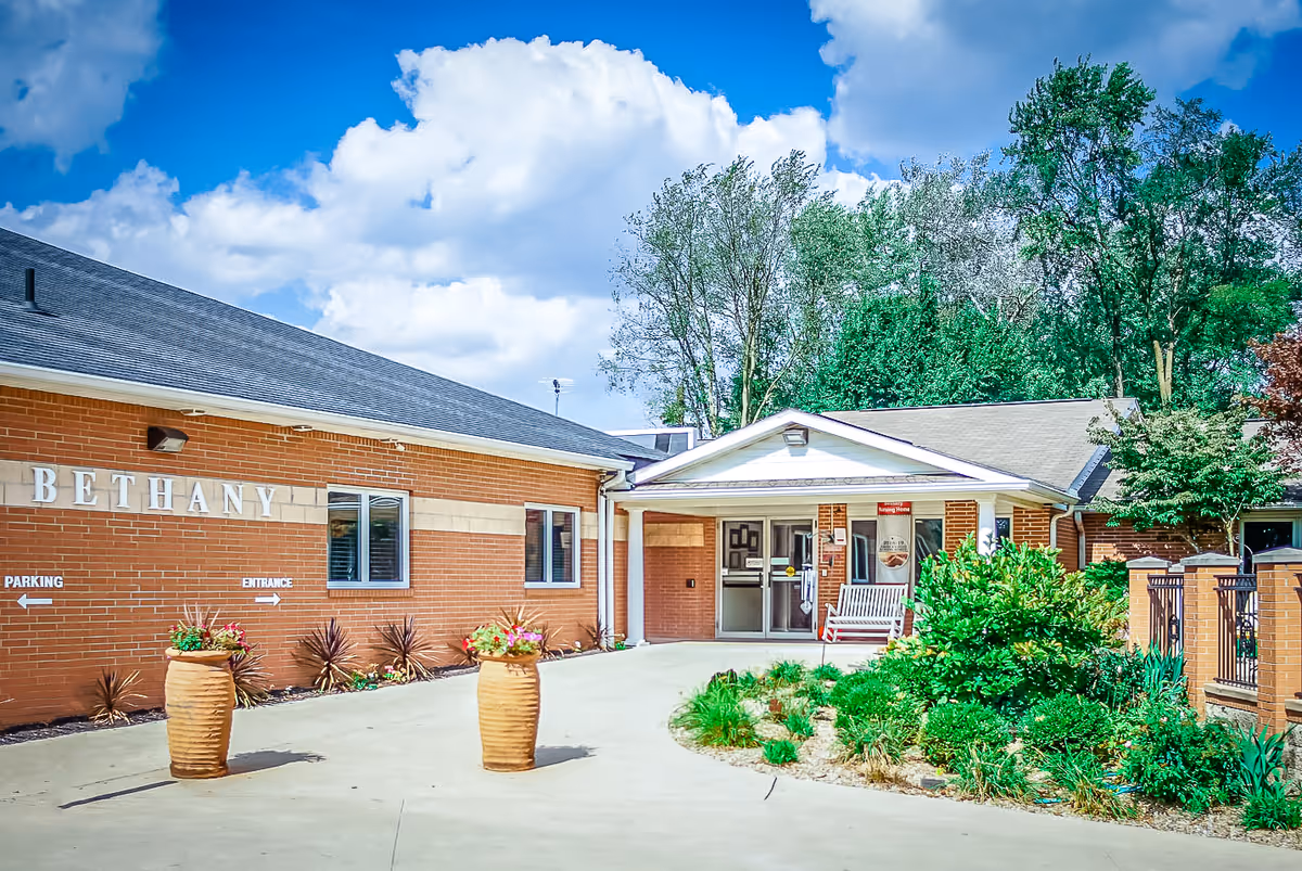 Exterior view of Bethany Nursing Home showing a brick building with a covered entrance, two large flower pots with plants, a bench near the entrance, and greenery including bushes and trees under a partly cloudy blue sky.