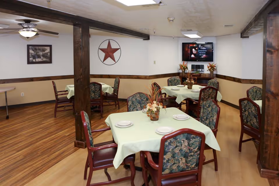 Dining room with several tables covered with light green tablecloths, each set with white plates and floral centerpieces. The room has wooden beams, floral upholstered chairs, a ceiling fan, a framed picture on the wall, and a TV mounted on the far wall displaying a music program.