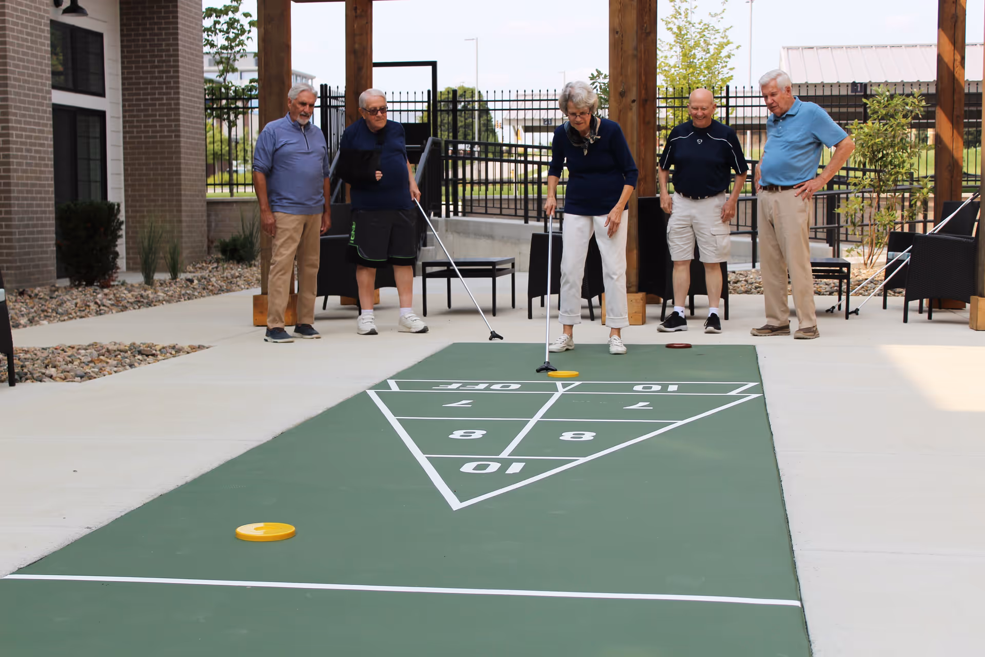 Five elderly people playing shuffleboard on an outdoor court at a senior living facility. They are standing on a concrete patio with a green shuffleboard court marked with numbers. The setting includes wooden posts, chairs, and a fenced area with some greenery in the background.