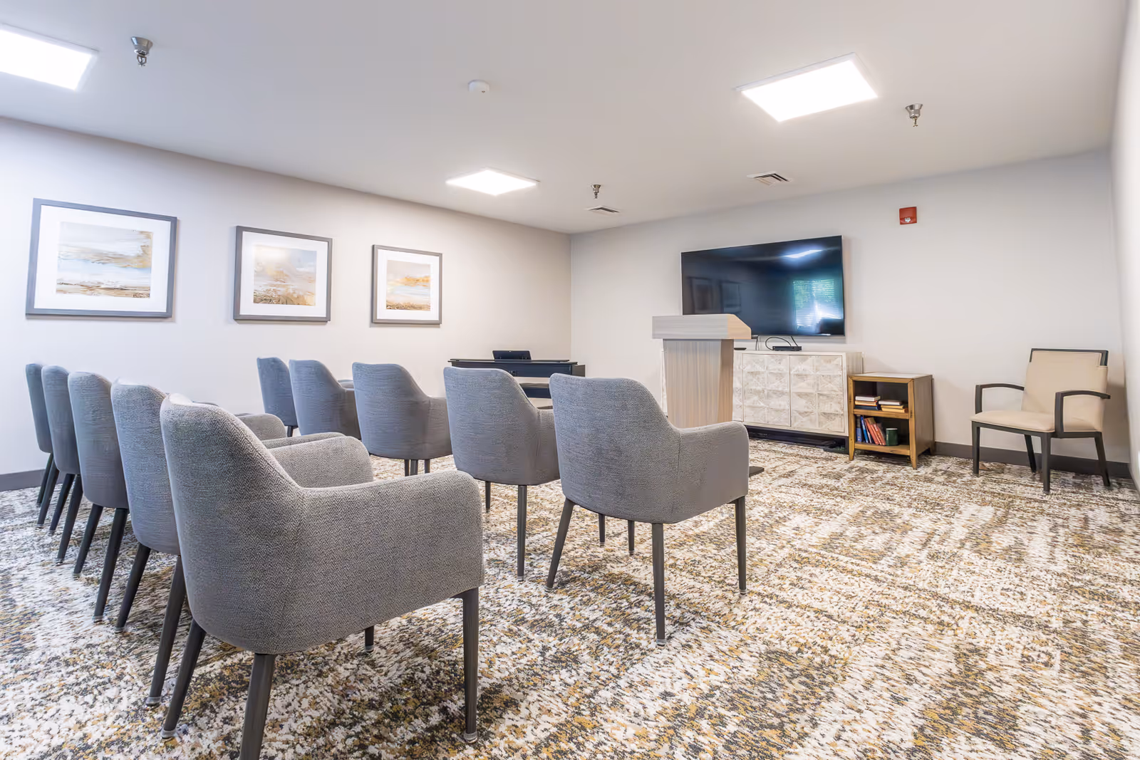 A carpeted community meeting room with rows of gray upholstered chairs facing a podium and a wall-mounted TV.