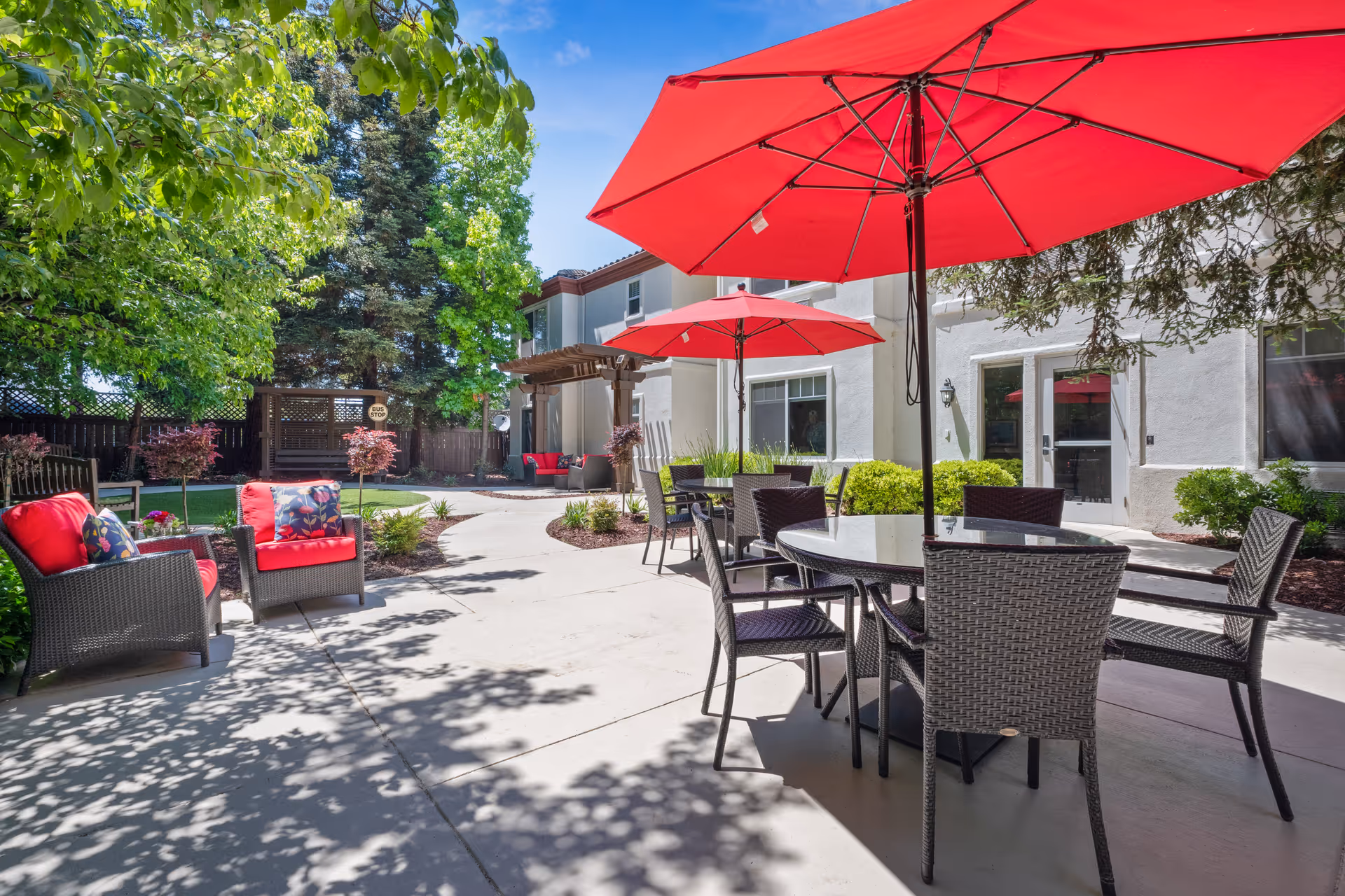 Outdoor patio area at Aegis Living Fremont with wicker chairs and tables under large red umbrellas. There are additional cushioned chairs with red cushions and floral pillows. The area is surrounded by green trees and shrubs, with a white building in the background and a wooden pergola seating area.