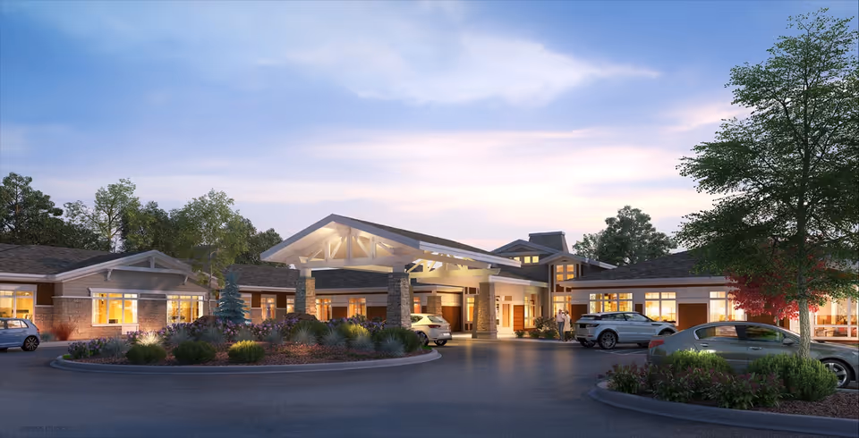 Exterior view of Bethesda Gardens Monument senior living facility at dusk, showing a large entrance with a covered driveway, stone pillars, landscaped roundabout with bushes and small trees, several parked cars, and warm interior lights visible through windows.