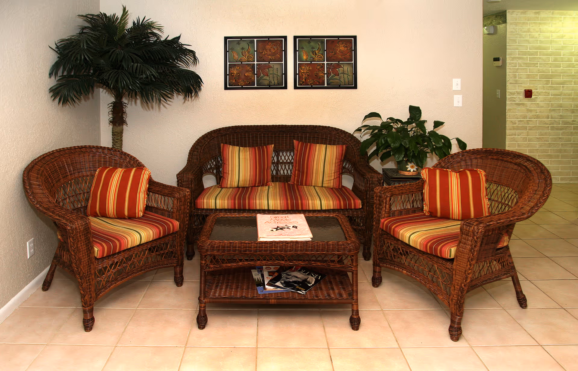 A cozy seating area with two wicker armchairs and a wicker loveseat, all with striped red, yellow, and orange cushions. A matching wicker coffee table with a glass top holds a book and magazines underneath. There are two framed floral artworks on the wall above the loveseat, a potted plant on a small table to the right, and a tall artificial palm tree in the corner to the left. The floor is tiled and the walls are light-colored.
