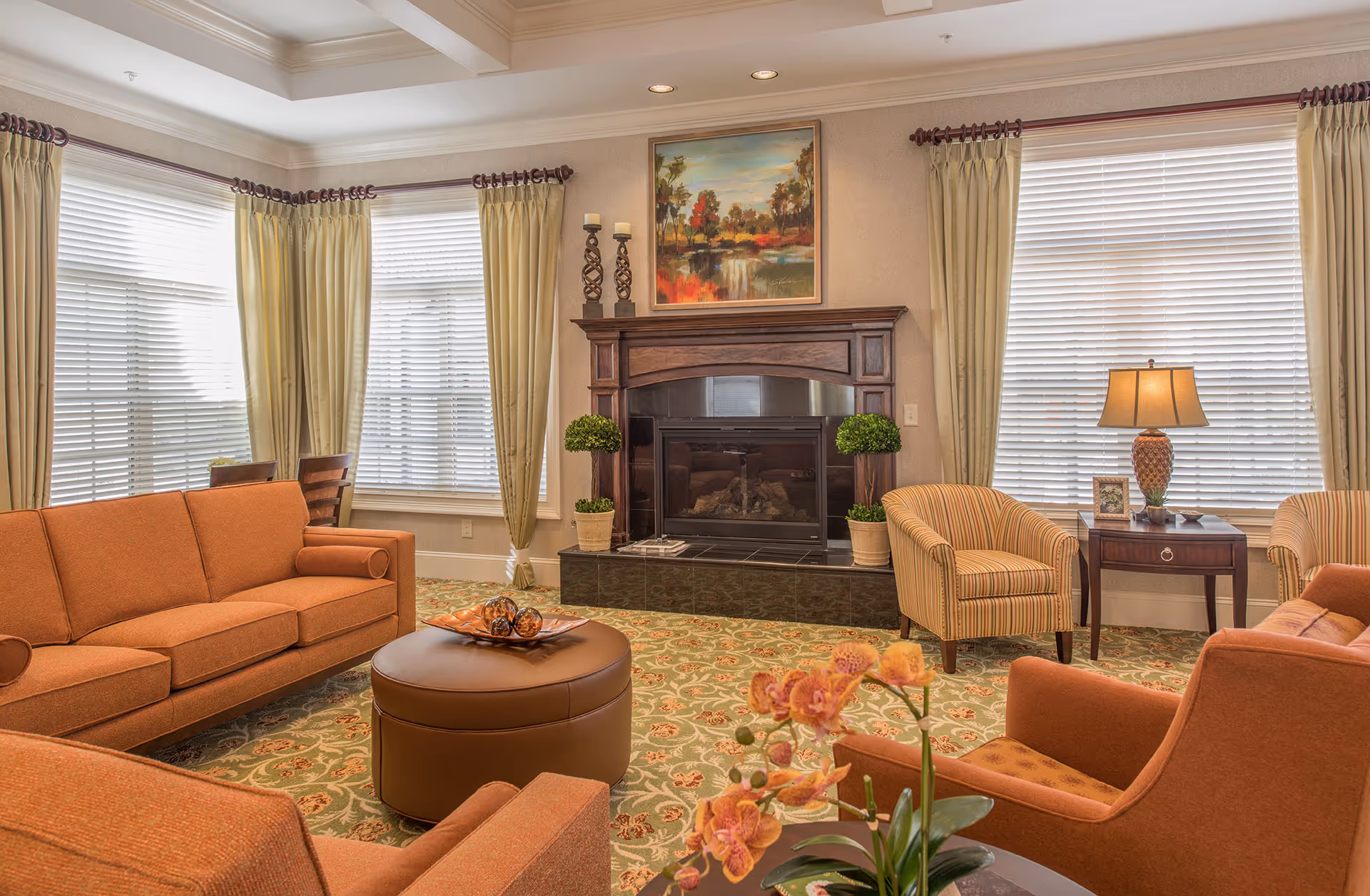 A cozy living room with orange upholstered sofas and armchairs arranged around a round brown ottoman. The room features large windows with cream-colored curtains, a fireplace with a dark wooden mantel, two potted plants on either side, and a colorful landscape painting above the fireplace. A side table with a lamp and framed photo is positioned near one of the armchairs. The floor is covered with a patterned carpet.