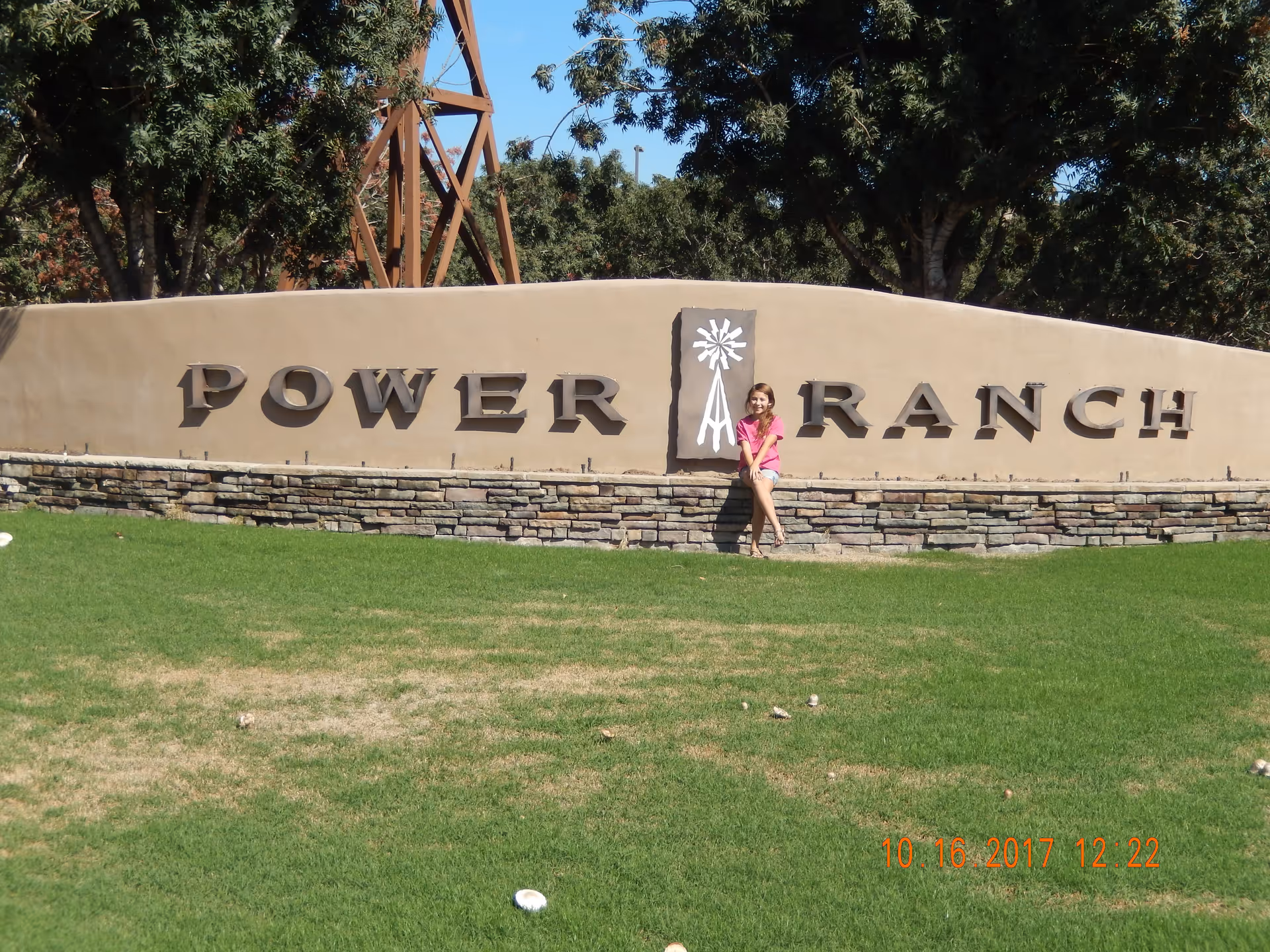 A young girl in a pink shirt and shorts sitting on a stone and stucco sign that reads 'POWER RANCH' with a windmill symbol in the center, surrounded by green grass and trees in the background under a clear blue sky.