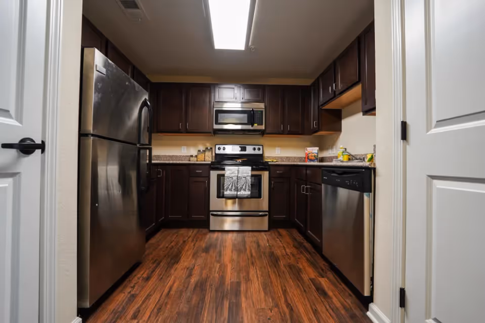 Modern kitchen with dark wood cabinets, stainless steel refrigerator, oven, microwave, and dishwasher. The kitchen has a wooden floor and bright overhead lighting.