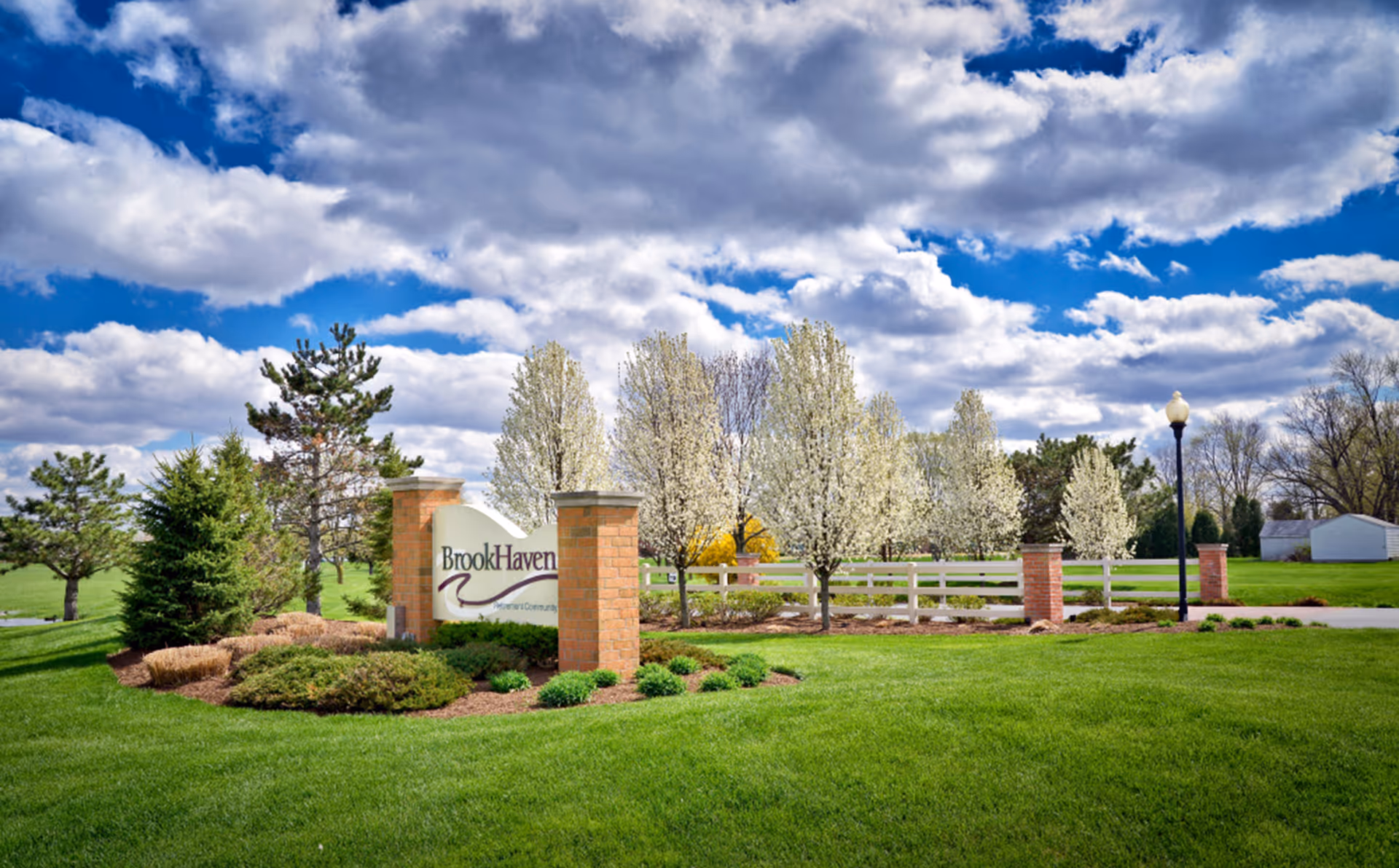 A landscaped outdoor area with green grass, flowering trees, and a brick sign that reads 'BrookHaven Retirement Community' under a partly cloudy blue sky.