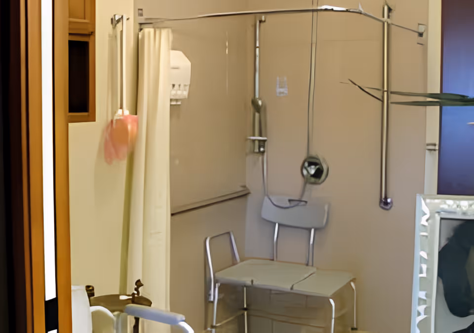 A bathroom with a shower area featuring a white shower curtain, a metal shower chair with a backrest, and grab bars on the wall for support. There is a soap or shampoo dispenser mounted on the wall inside the shower area.