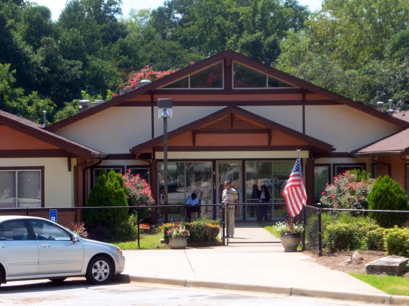 Front exterior view of a single-story building with a triangular roof, surrounded by greenery and flowering bushes. There is a sidewalk leading to the entrance, where a few people are standing near the glass doors. An American flag is displayed near the entrance, and a silver car is parked in front.
