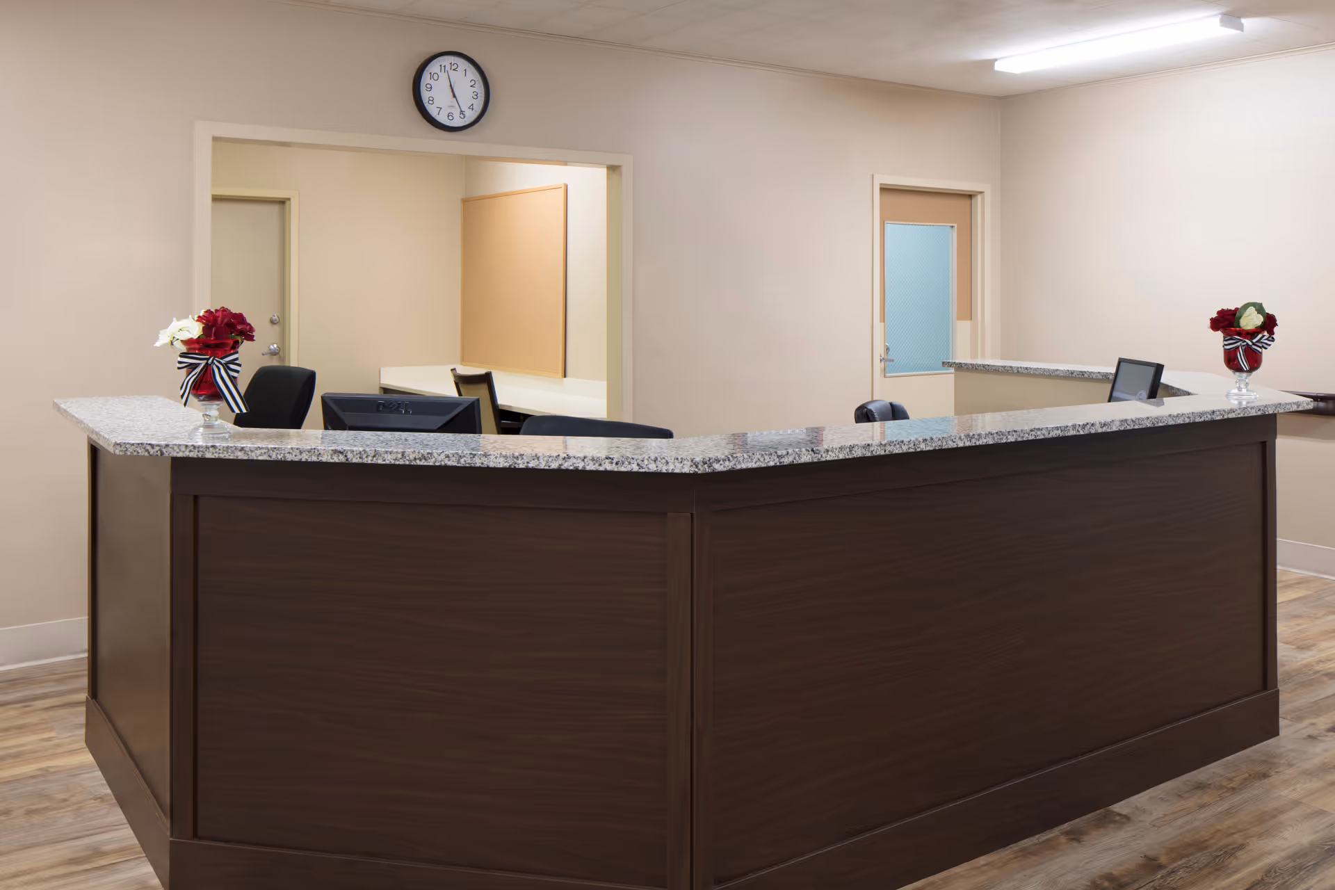 Reception desk area with a dark wood counter and granite top, two computer monitors, two vases with red and white flowers tied with black and white striped ribbons, beige walls, a clock showing 7:05, and two doors in the background.