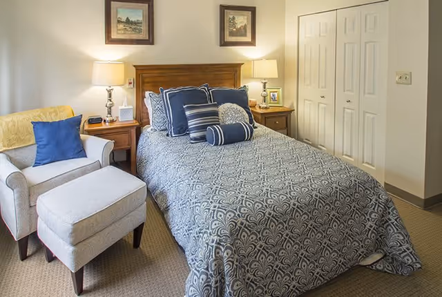 A cozy bedroom featuring a bed with a patterned blue and white bedspread and multiple decorative pillows. On either side of the bed are wooden nightstands with lamps, a clock, and a framed photo. To the left of the bed is a cushioned armchair with a matching ottoman and a blue throw pillow. The room has beige walls, two framed pictures above the bed, and a double-door closet on the right side.