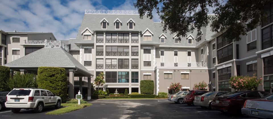 Front exterior of a multi-story senior living facility with a porte-cochere, landscaped entrance, and cars parked in the driveway.