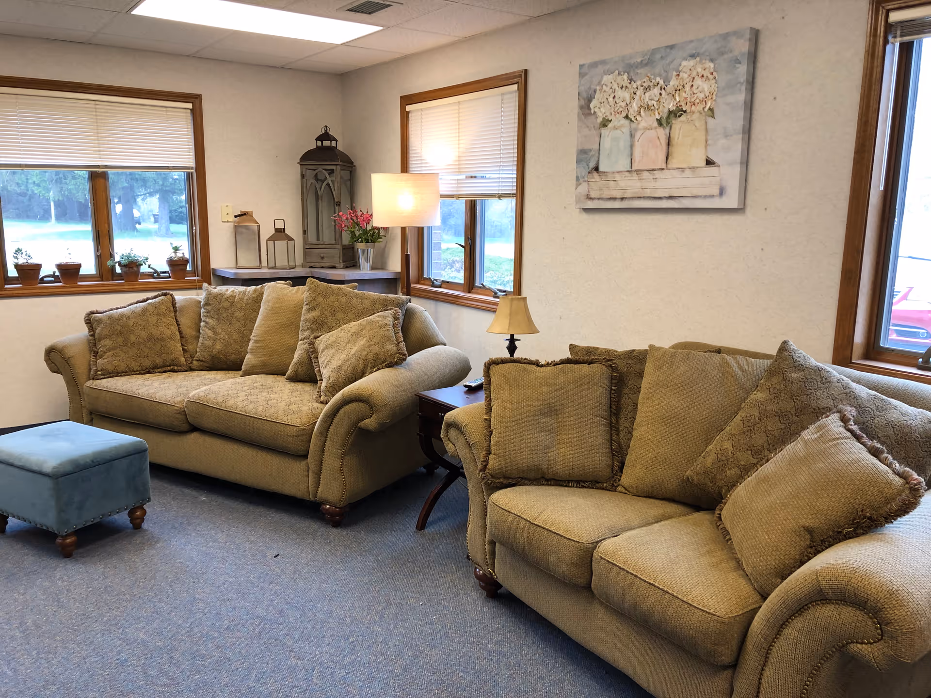 A cozy living room with two beige sofas adorned with multiple cushions, a small blue ottoman, a wooden side table with a lamp, and decorative items including potted plants on the windowsill and lanterns on a corner table. The room has three windows with blinds partially drawn and a painting of flowers on the wall.