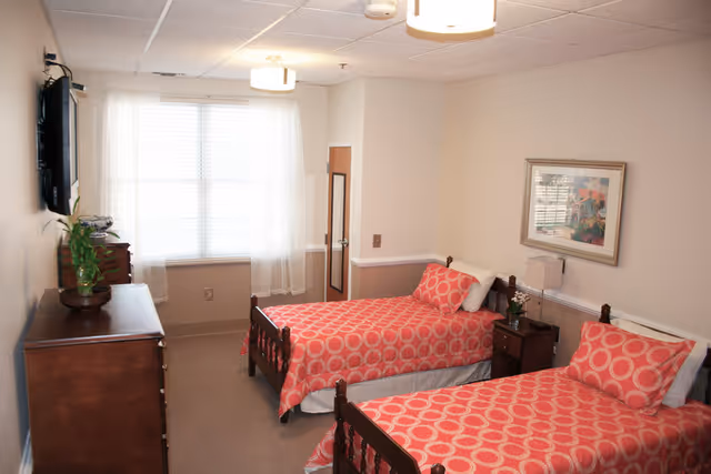 Two twin beds with red patterned bedspreads in a well-lit senior living bedroom with a dresser, wall-mounted TV, and a window.