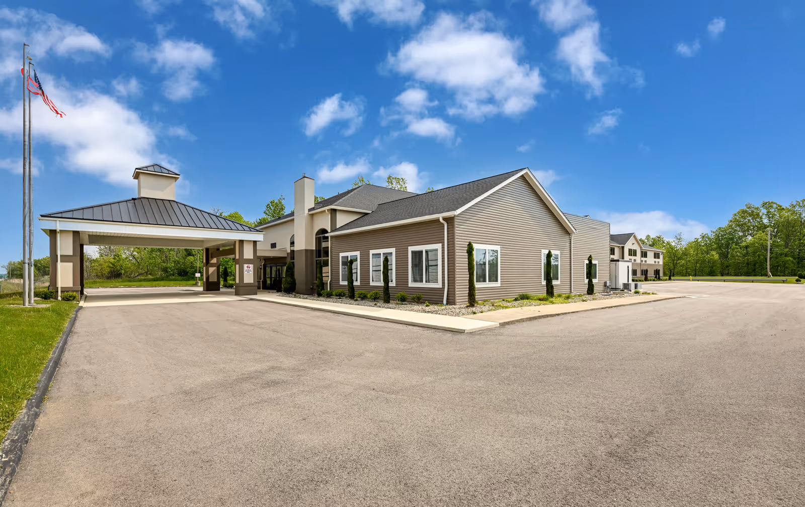 Exterior view of Hines Hill Senior Living facility showing a large paved driveway, a covered entrance, and a two-story building with beige siding and white trim under a blue sky with scattered clouds. An American flag flies on a flagpole to the left.