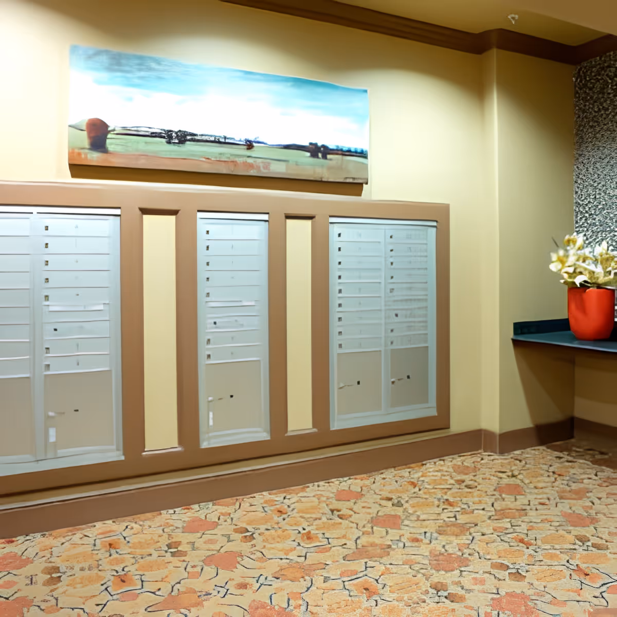 Interior view of a mailroom area with multiple mailboxes mounted on a beige wall. Above the mailboxes is a landscape painting with blue sky and green fields. To the right, there is a countertop with a red vase holding white flowers. The floor has a patterned carpet with earth tones.
