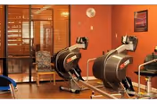 Indoor exercise room with two stationary exercise bikes positioned on a wooden floor. There is a chair with a floral cushion near a window with wooden blinds, and a framed picture on the orange wall.