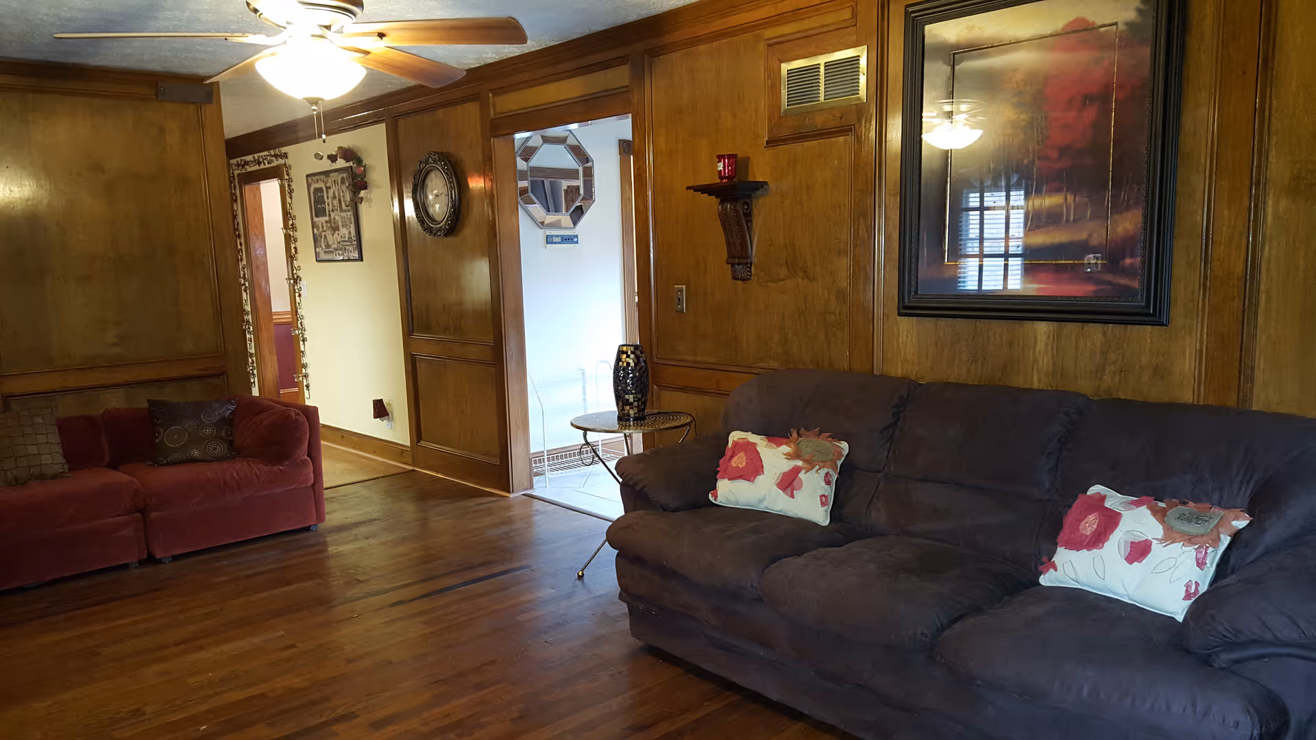 A cozy living room with wooden paneled walls and hardwood floors. The room features a dark brown sofa with floral pillows on the right and a red loveseat with a decorative pillow on the left. A ceiling fan with a light is mounted on the ceiling. There is a framed painting of trees with red foliage on the wall above the sofa, and a small round side table with a decorative vase next to it. The room opens into a hallway with additional wall decor including a clock and framed pictures.