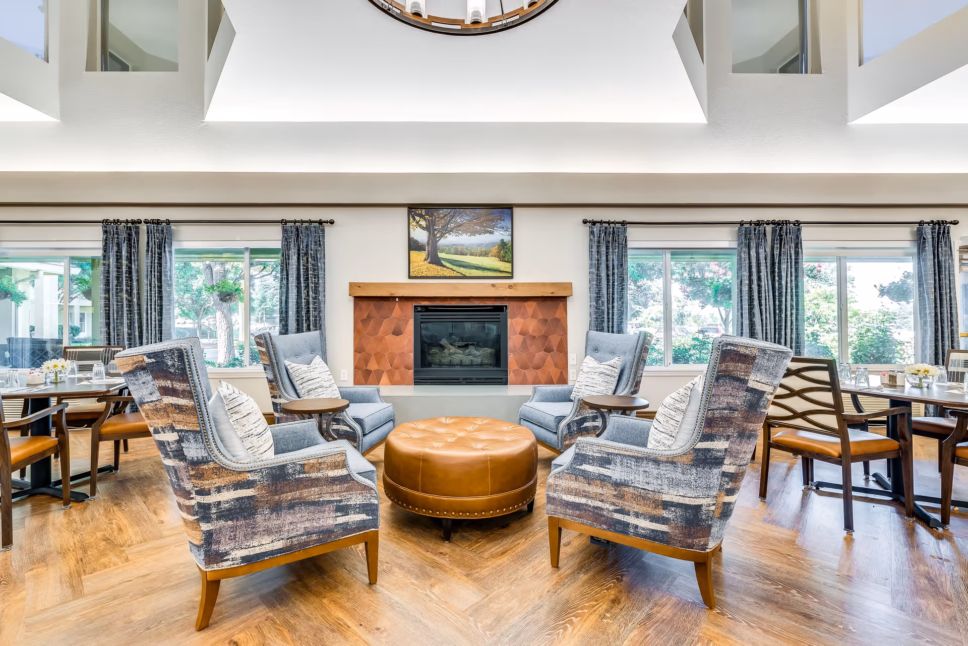 A bright and inviting senior living common area featuring four patterned armchairs arranged around a round, tufted leather ottoman in front of a modern fireplace with a wooden mantel. Large windows with dark curtains let in natural light and show greenery outside. Dining tables and chairs are visible on either side of the seating area. The room has wood flooring and a high ceiling with a circular architectural feature above.