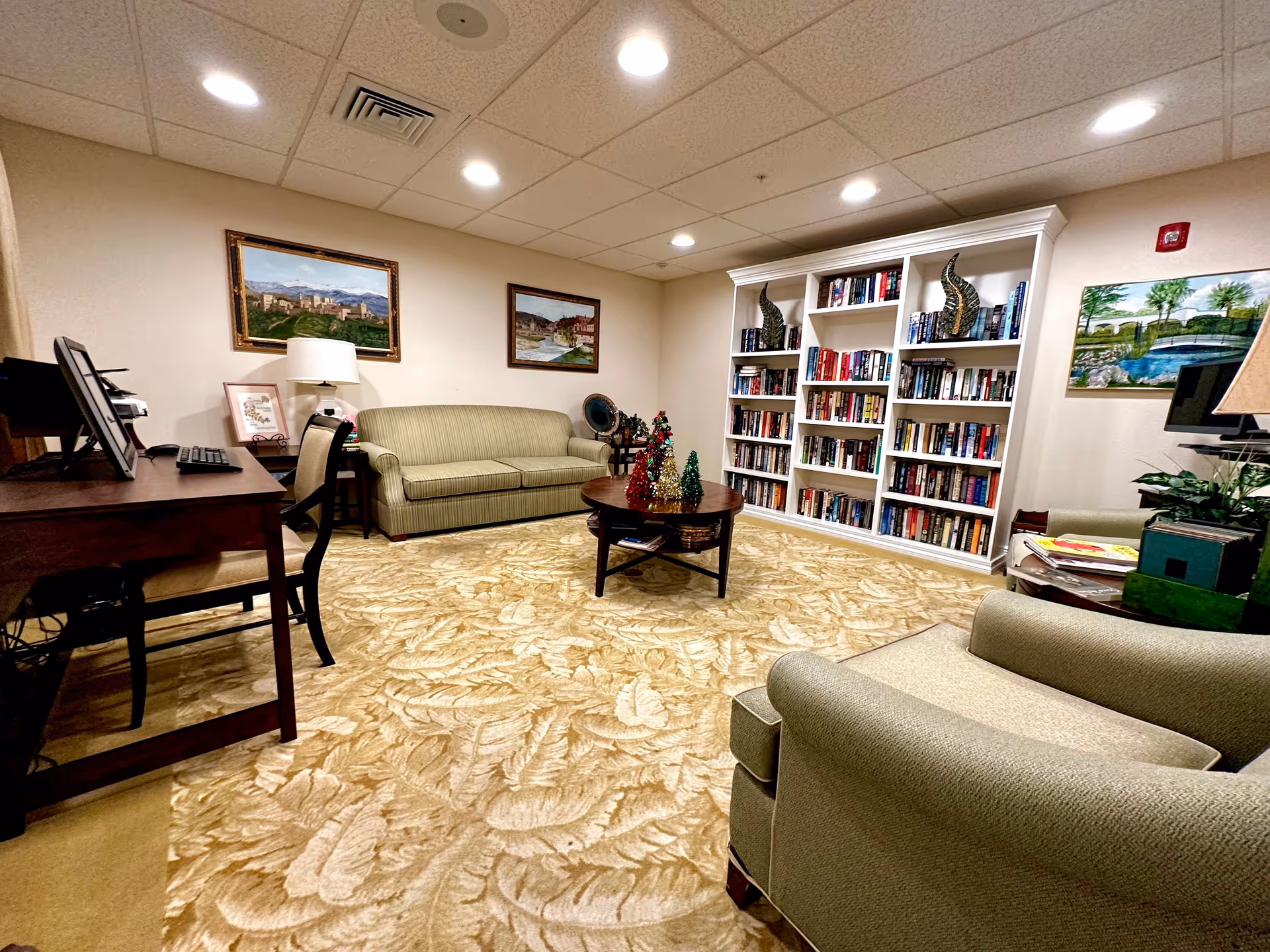 A cozy senior living common area with a green striped sofa, matching armchair, wooden coffee table decorated with small colorful Christmas trees, a large white bookshelf filled with books, a wooden desk with a computer, and framed landscape paintings on the walls. The room has a beige patterned carpet and recessed ceiling lights.