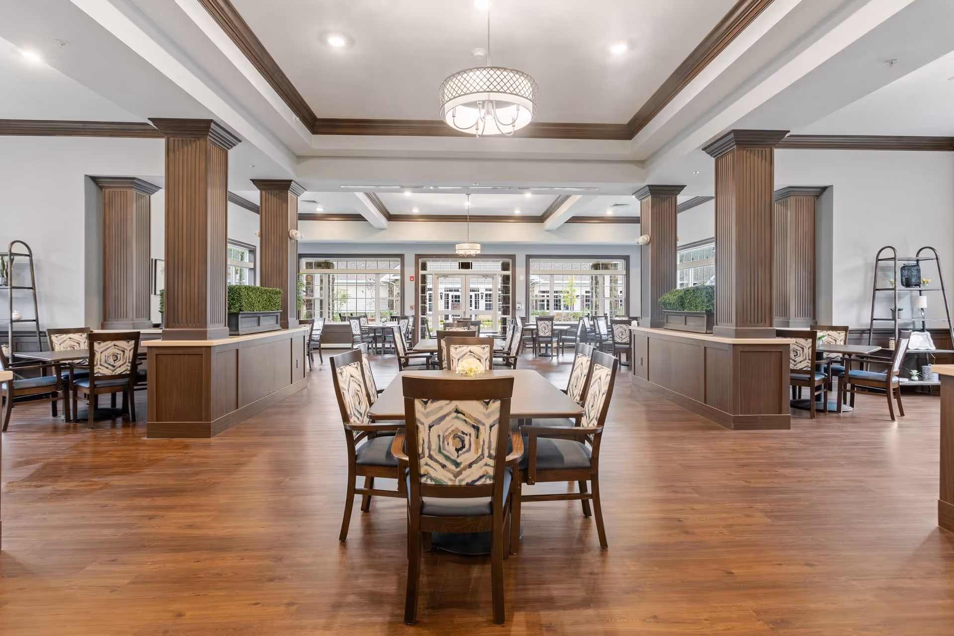 Spacious dining room with wooden floors, multiple tables and chairs with patterned upholstery, large windows letting in natural light, decorative wooden columns, and modern ceiling lights.