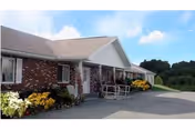 Single-story brick assisted-living building with a covered entrance, accessibility ramp, shrubs and a paved driveway under a partly cloudy sky.