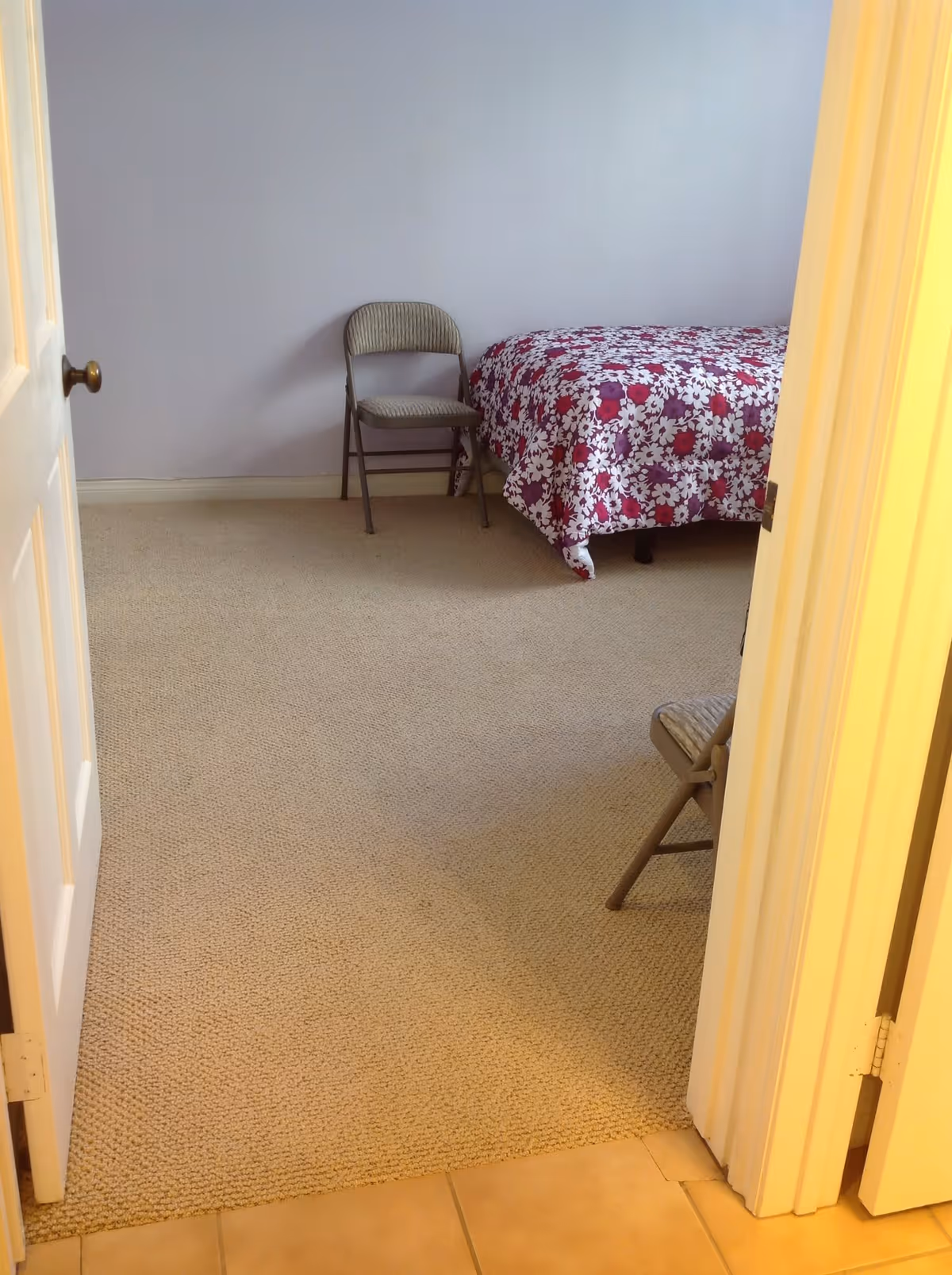 View through an open doorway into a bedroom with beige carpet, a bed with a floral red and white bedspread, and two folding chairs, one against the wall and one partially visible near the door.