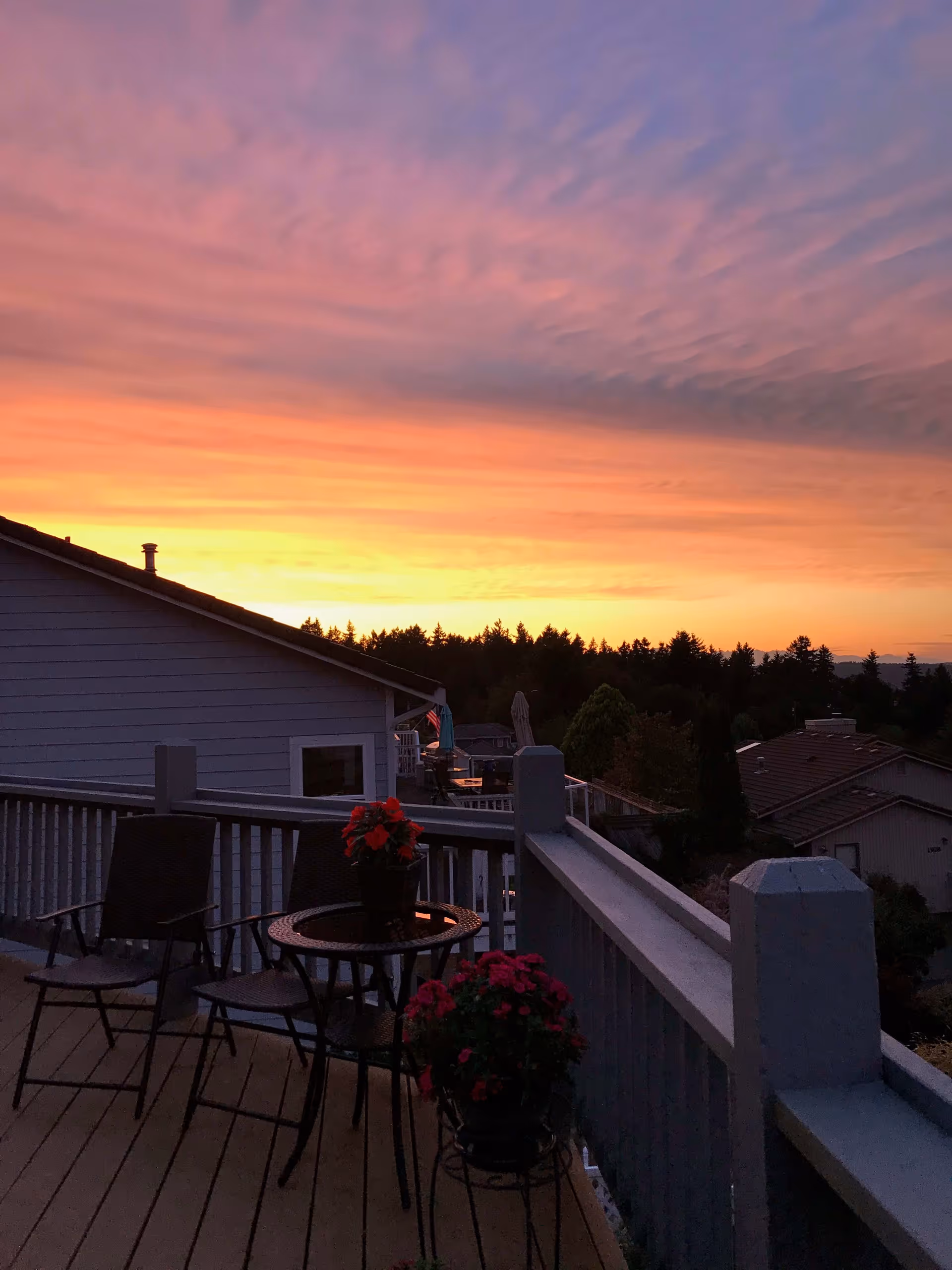 A balcony at sunset with two chairs and a small round table holding a potted plant with red flowers. Another pot with red flowers is on a stand nearby. The balcony overlooks neighboring houses and trees under a colorful sky with shades of orange, pink, and purple.