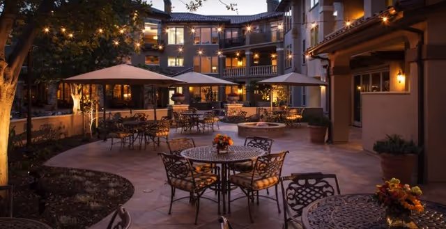 Outdoor patio area at dusk with multiple round metal tables and chairs, some tables decorated with flower arrangements. Large umbrellas provide shade over some tables. String lights are hung above, creating a warm and inviting atmosphere. The multi-story building of the Villagio at Capriana Memory Care Community is visible in the background with lit windows.