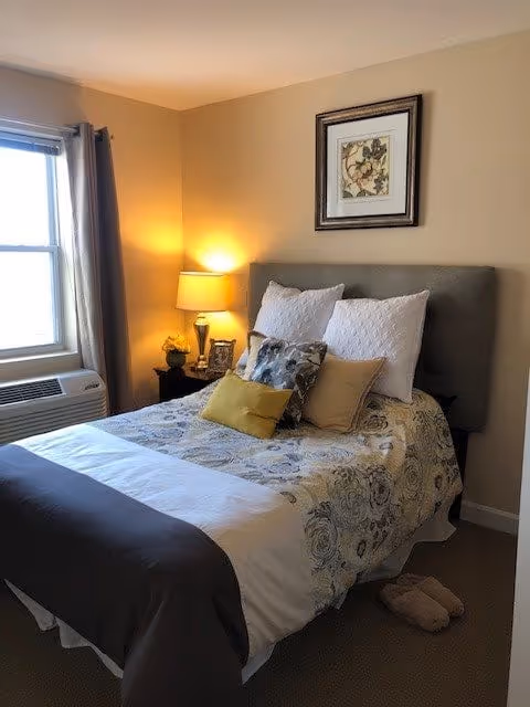 A cozy bedroom with a neatly made bed featuring a floral-patterned bedspread, multiple pillows, and a gray upholstered headboard. A bedside table with a lamp, a small plant, and a framed photo is next to the bed. A window with curtains and an air conditioning unit is on the left wall. A framed floral artwork hangs above the headboard, and a pair of slippers is on the floor near the foot of the bed.
