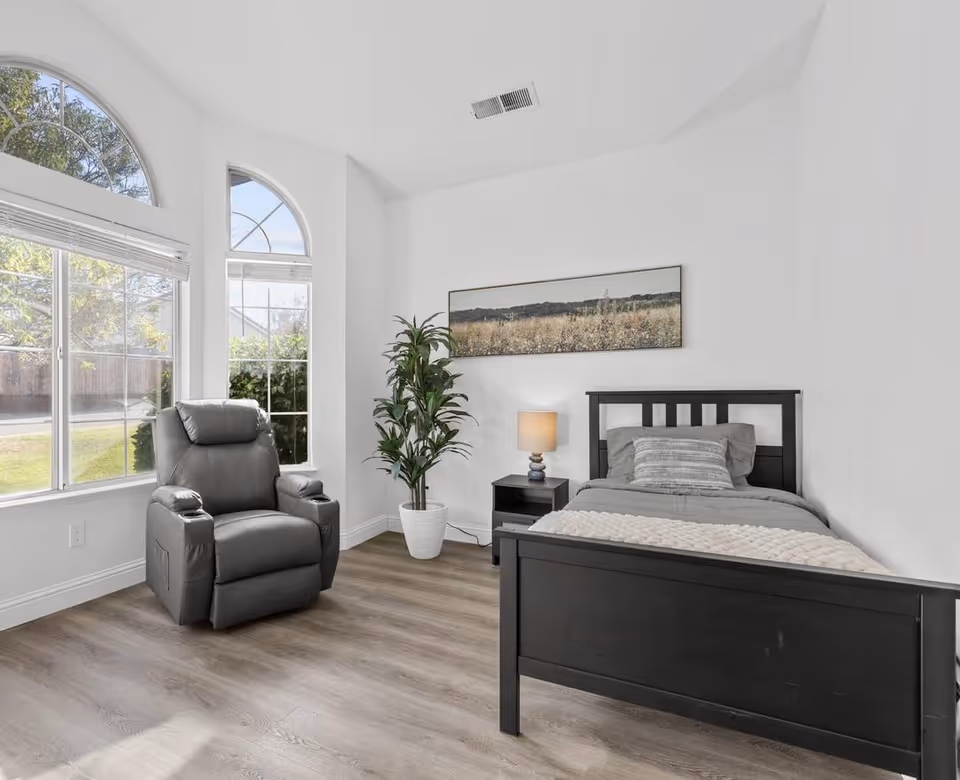 Bright single bedroom with a black bed and nightstand, lamp, a gray recliner by large arched windows, and a potted plant.
