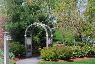 A garden area with a paved pathway leading through a metal archway surrounded by green bushes, trees, and various plants under a clear sky.