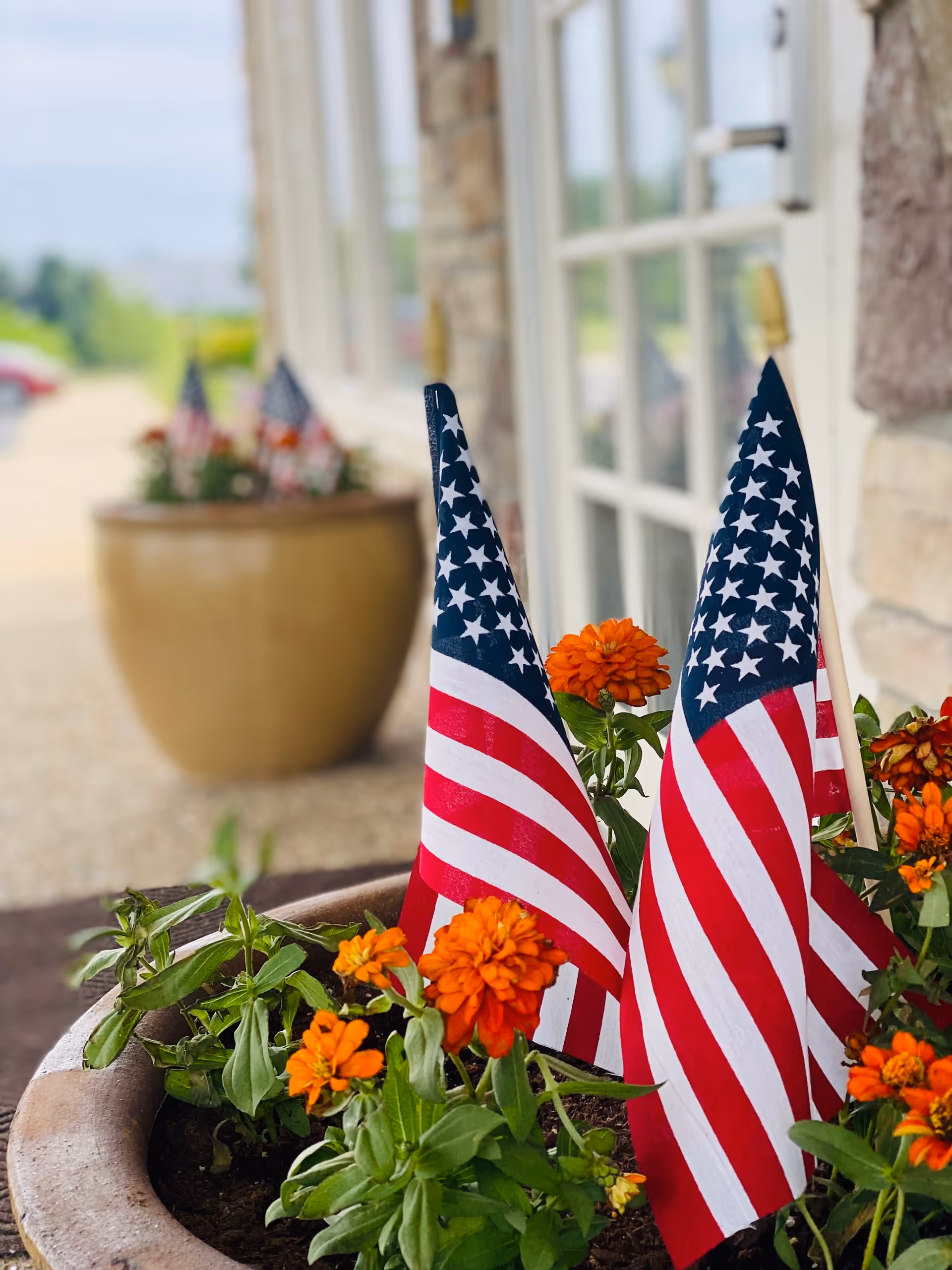 Close-up view of a planter with orange flowers and two small American flags placed in the soil. In the background, there is another planter with similar flowers and flags, and a building with large windows and stone walls.