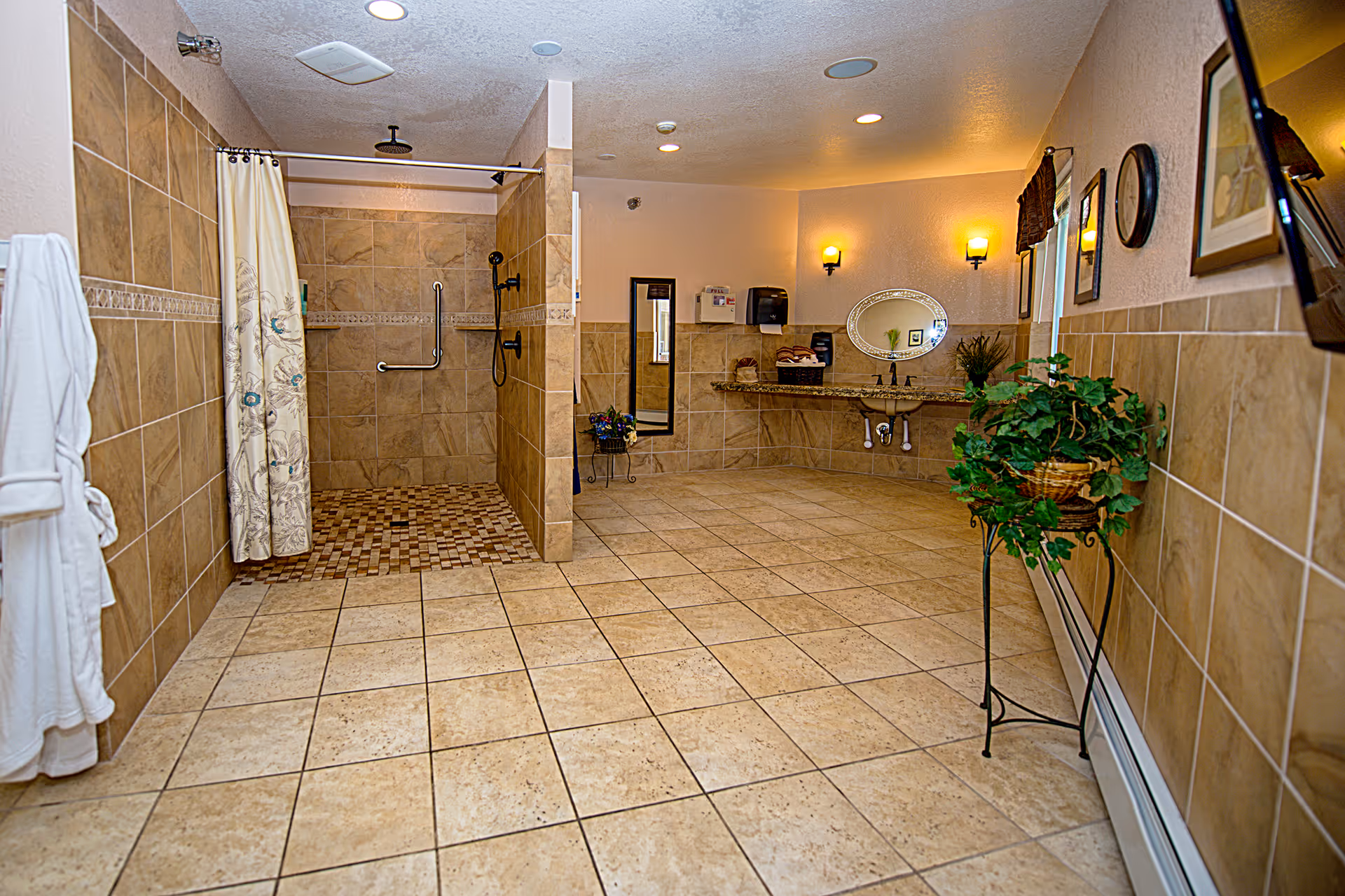 Spacious bathroom with beige tiled floors and walls, featuring a walk-in shower with a curtain and grab bars, a wall-mounted showerhead, a large mirror above a granite countertop with a sink, two wall sconces providing warm lighting, a full-length mirror, a white bathrobe hanging on the wall, and a potted plant on a stand.