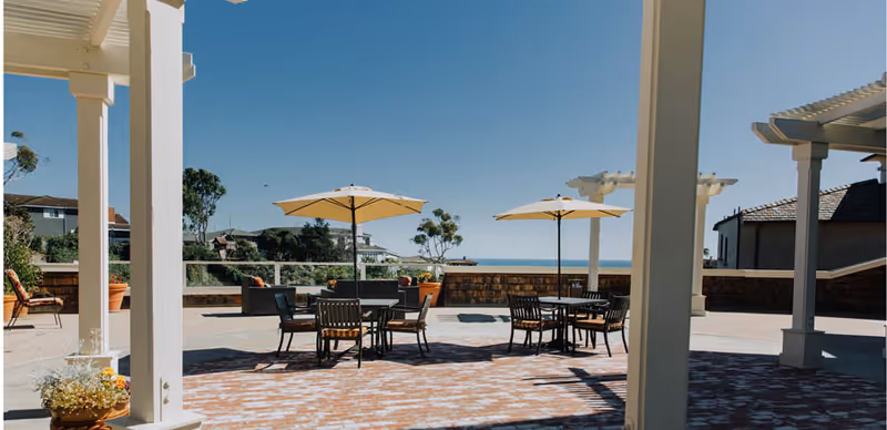 Outdoor patio area with several tables and chairs under large beige umbrellas, surrounded by white pergolas and potted plants, with a clear blue sky and distant trees and houses in the background.