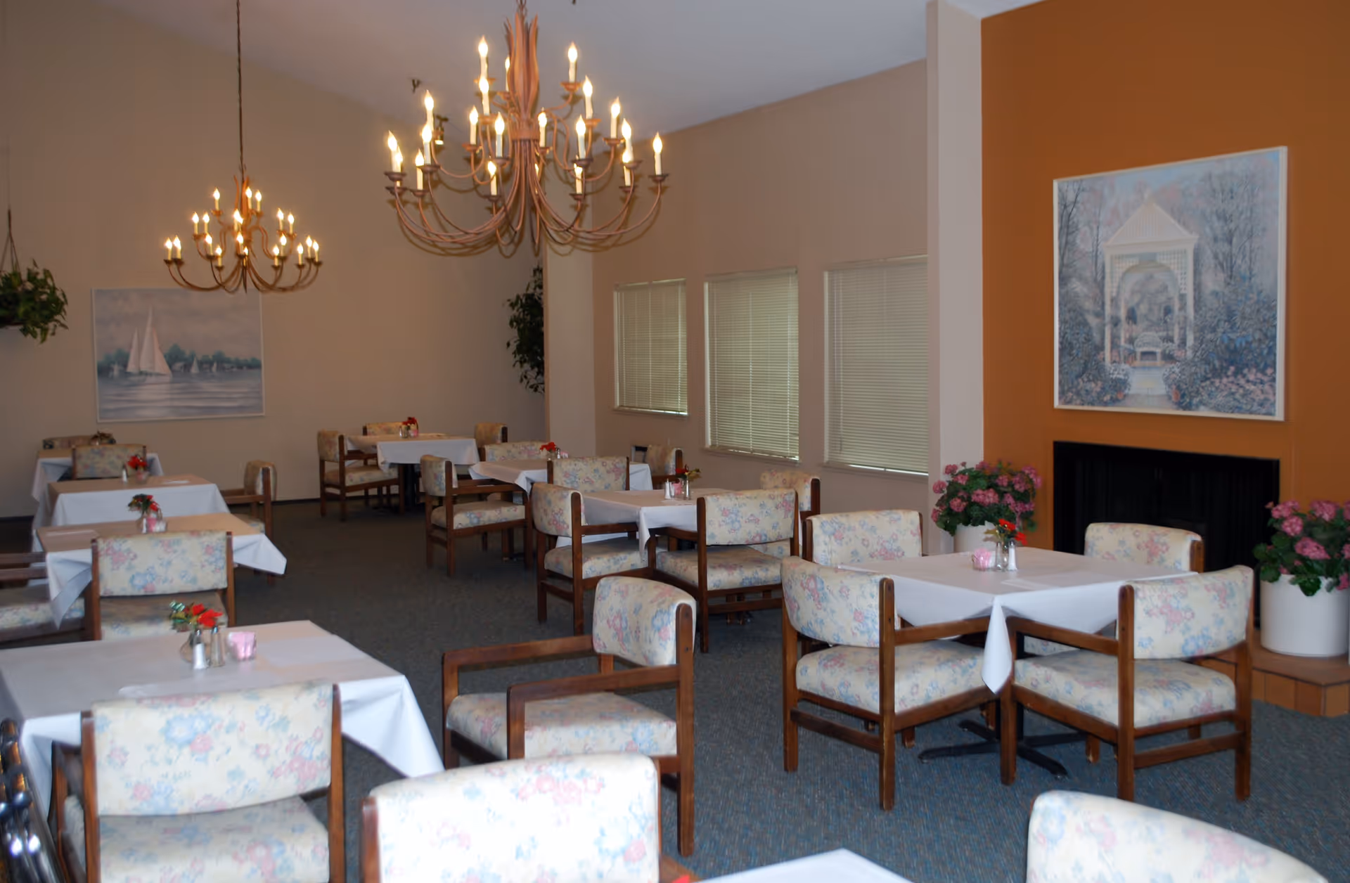 A dining room with multiple tables covered with white tablecloths, surrounded by floral upholstered chairs. The room features two large chandeliers, several paintings on the walls, and potted plants, including flowers on the tables and near a fireplace with an orange accent wall.