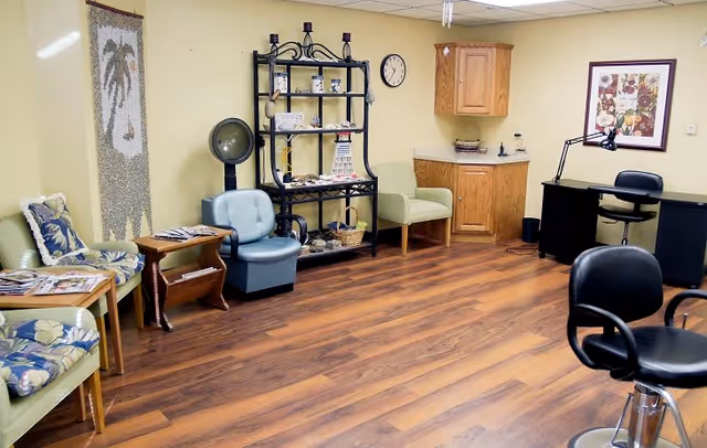 A cozy interior room with wooden flooring featuring several chairs including cushioned armchairs and swivel chairs. There is a black metal shelf with decorative items, a small wooden side table with magazines, a wall clock, a framed floral picture, and a wooden cabinet with a countertop. The walls are painted light yellow.
