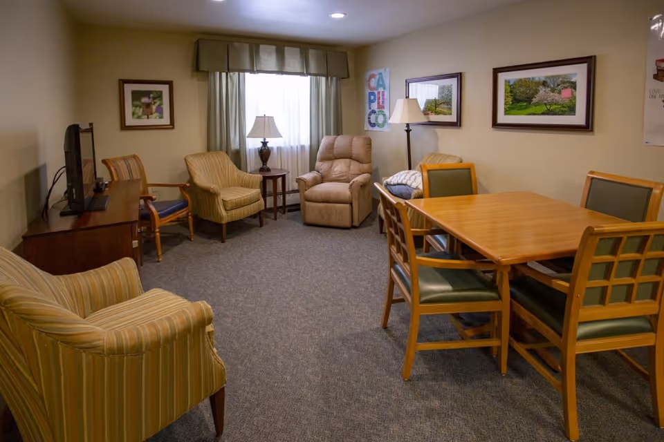A cozy living room in an assisted living facility with a beige recliner, several armchairs, a wooden table with four chairs, two table lamps, framed pictures on the walls, and a window with curtains letting in natural light.