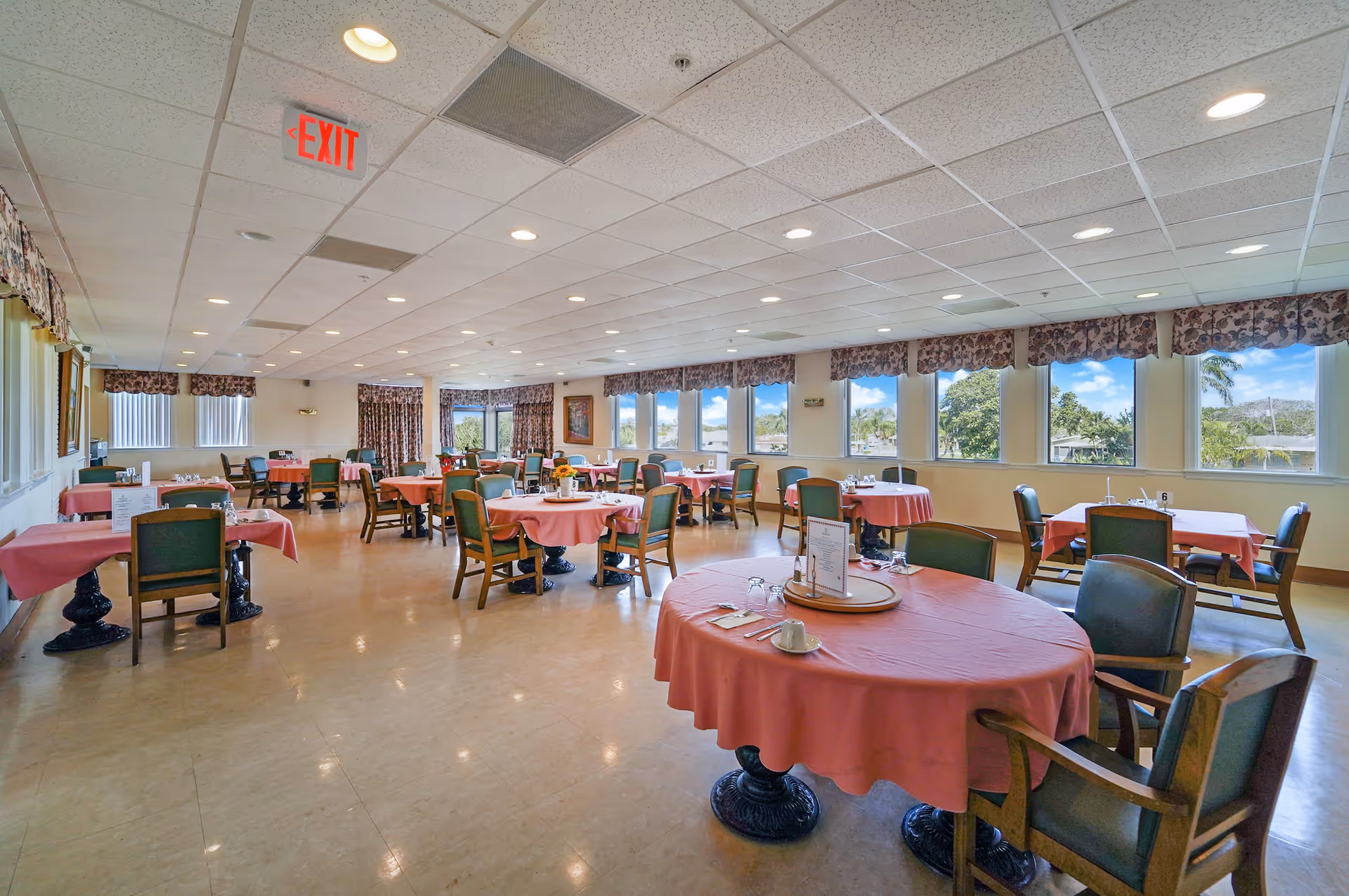 A spacious dining room with multiple round tables covered with pink tablecloths, each surrounded by wooden chairs with green cushions. The room has large windows along one wall letting in natural light and offering a view of trees and a blue sky. The ceiling is fitted with recessed lighting and an exit sign is visible on the ceiling.