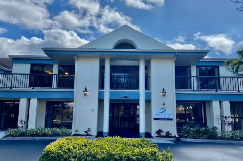 Front exterior view of a two-story building with white walls and blue accents under a partly cloudy sky. The building has a central entrance with four white columns and a balcony above. There is a sign near the entrance that reads 'Springwood Court'.
