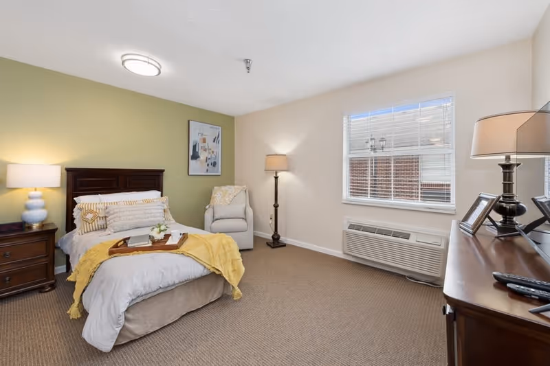 A cozy bedroom with a bed featuring white and yellow bedding, a wooden nightstand with a lamp and clock, a comfortable armchair with a throw blanket, a floor lamp, a window with blinds, and a wooden dresser with a lamp and picture frames.