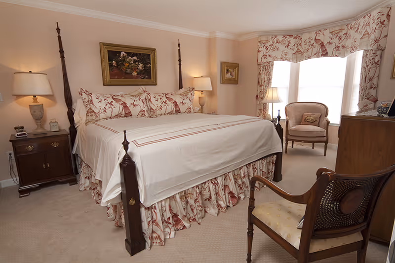 Traditional bedroom with a four-poster bed, bedside tables and lamps, upholstered chairs, and a bay window with patterned curtains.