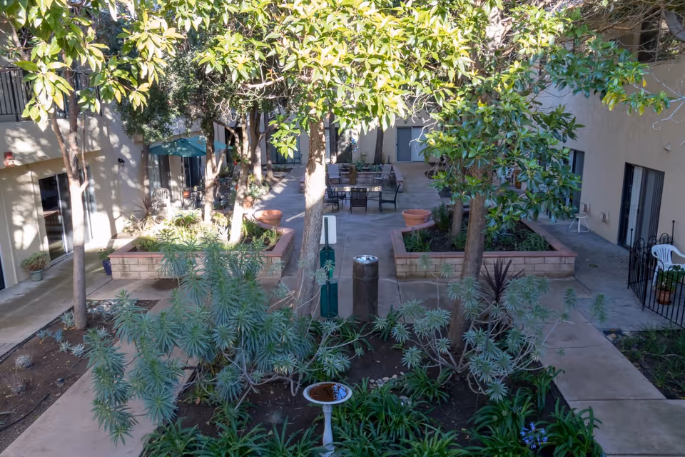 Outdoor courtyard area at Casa De Flores featuring raised garden beds with various plants and trees, surrounded by a paved walkway. There are patio tables and chairs under umbrellas in the background, with building entrances visible on both sides.