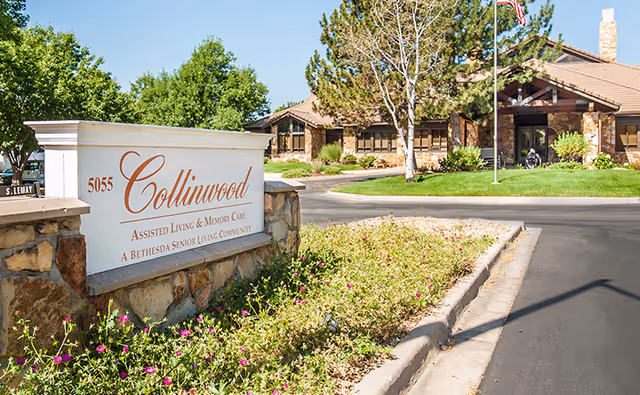Entrance sign for Collinwood Assisted Living & Memory Care with a stone base and white panel displaying the facility name and address. Behind the sign is a driveway leading to a single-story building surrounded by trees and greenery under a clear blue sky.