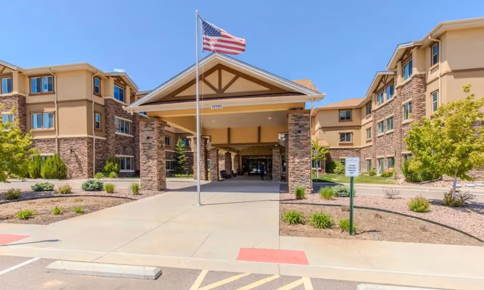 Front exterior view of Pine Grove Crossing facility with a covered entrance supported by stone pillars, an American flag on a flagpole, and landscaped areas with small trees and shrubs under a clear blue sky.