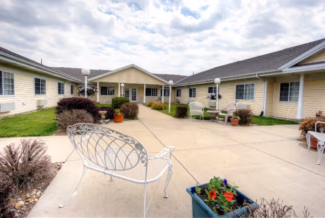 Outdoor courtyard with paved walkways, white metal benches, potted plants, and a single-story yellow senior living building under a cloudy sky.