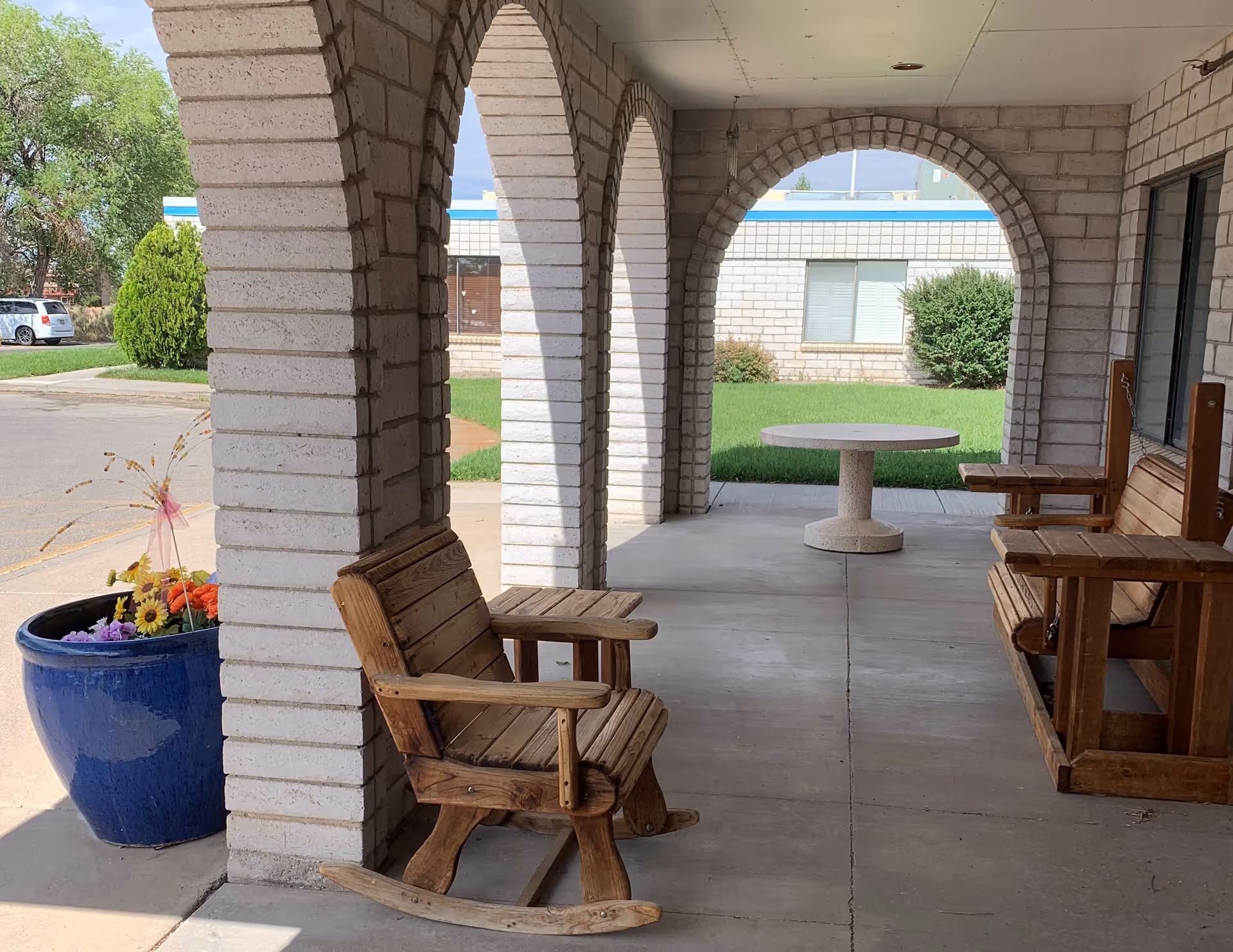 Covered outdoor patio area with white brick arches, wooden rocking chairs, a wooden bench swing, a round stone table, and a large blue planter with colorful flowers. Green grass and a building with windows are visible in the background.