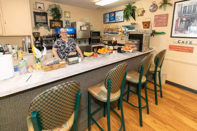 A woman stands behind a counter in a small café area with four high chairs in front. The counter has various items including fruit, cookies, and a Pepsi sign. The walls are decorated with framed pictures, plants, and signs, one of which reads 'CAFÉ'.